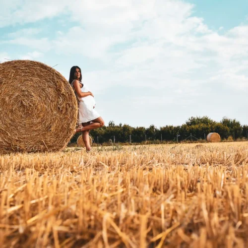 Una donna incinta in un vestito bianco elegante posa accanto a un rotolo di fieno in un campo dorato sotto un cielo azzurro, ideale per servizi fotografici di moda e book fotografici naturali.