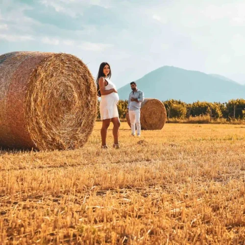 Una coppia in attesa dolcemente davanti a un rotolo di fieno in un campo, con uno sfondo di colline, perfetto per servizi fotografici di moda e book fotografici emozionali.