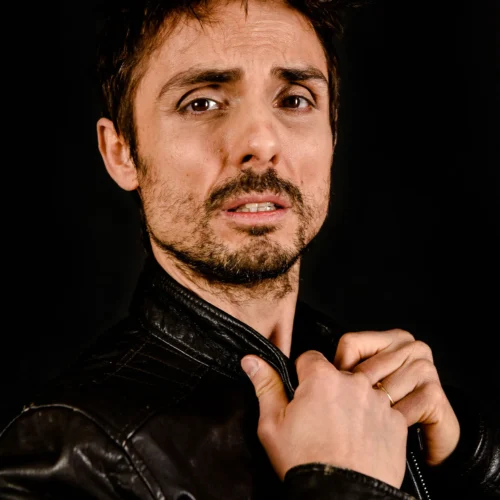 Scatto da servizi fotografici di moda e book fotografici che mostra un uomo in giacca di pelle nera, con le mani vicino al viso, su sfondo scuro, con un'espressione intensa.