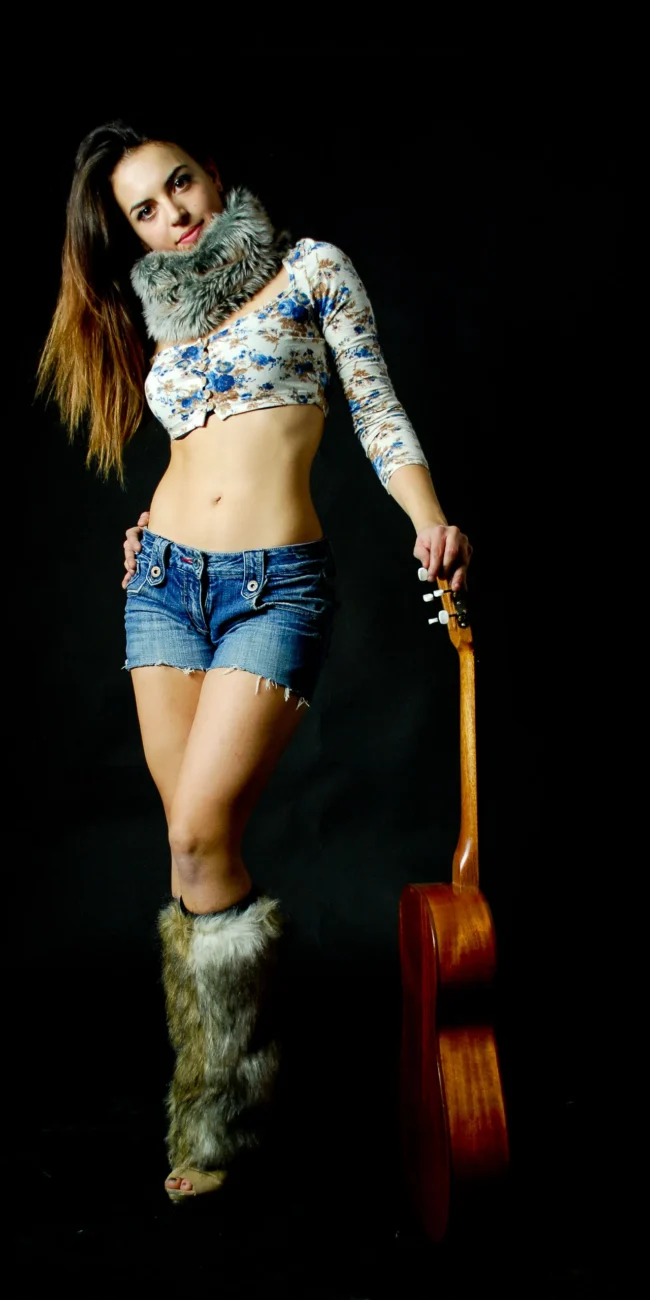 Modella in piedi con jeans e stivali di pelliccia, holding una chitarra, scatto di moda su sfondo nero per un book fotografico. (book fotografici, servizi fotografici di moda)
