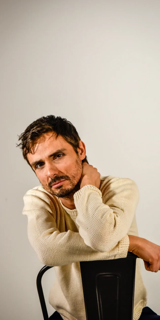 Immagine da servizi fotografici di moda e book fotografici che ritrae un uomo in maglione beige seduto su una sedia nera, con una mano sul viso, su sfondo chiaro.