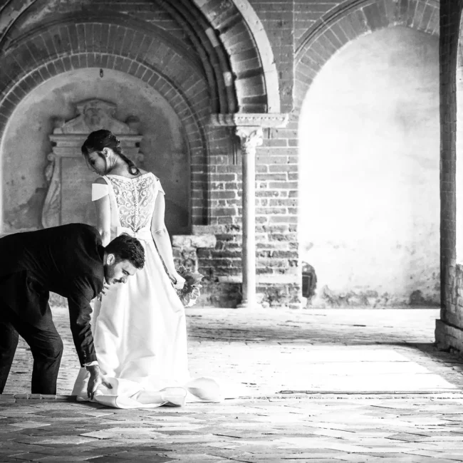 Immagine in bianco e nero di un reportage di matrimonio, con uno sposo che sistema il vestito della sposa in un chiostro con archi e colonne antiche.