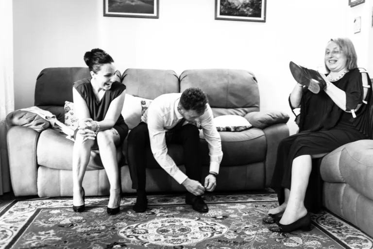 Reportage di matrimonio in bianco e nero, scena di una persona che allaccia una scarpa mentre altri osservano dal divano, con un'atmosfera intima.