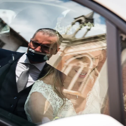 Sposa in abito bianco e uomo in abito scuro all'interno di un'auto, riflesso di un edificio sullo sfondo, foto matrimoniali, reportage di matrimonio.