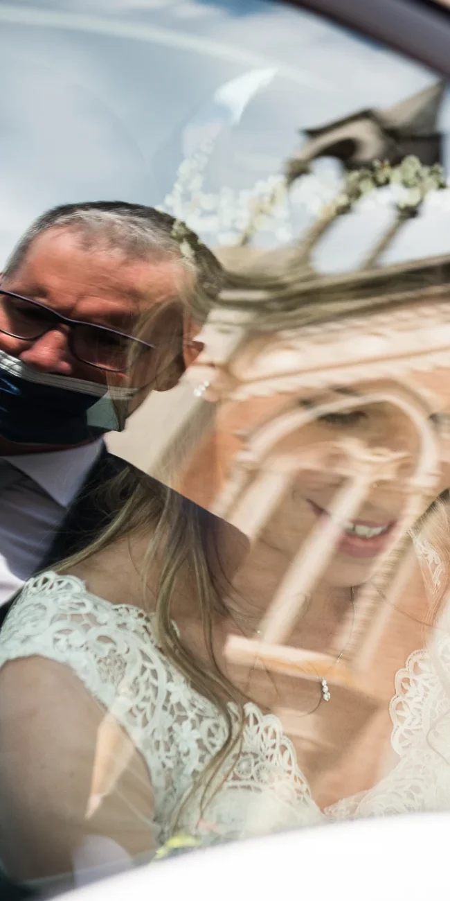 Sposa in abito bianco e uomo in abito scuro all'interno di un'auto, riflesso di un edificio sullo sfondo, foto matrimoniali, reportage di matrimonio.