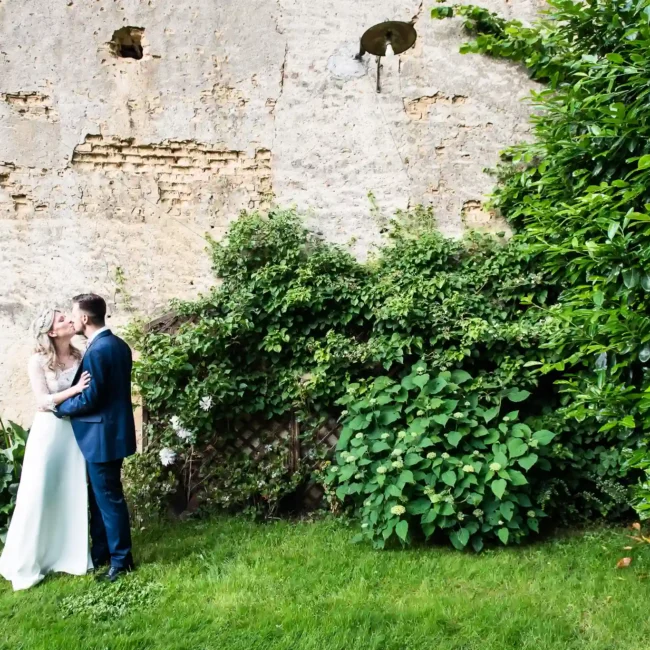 Foto matrimoniale di una coppia di sposi che si bacia su un prato con un muro di pietra e vegetazione sullo sfondo, parte di un reportage di matrimonio.