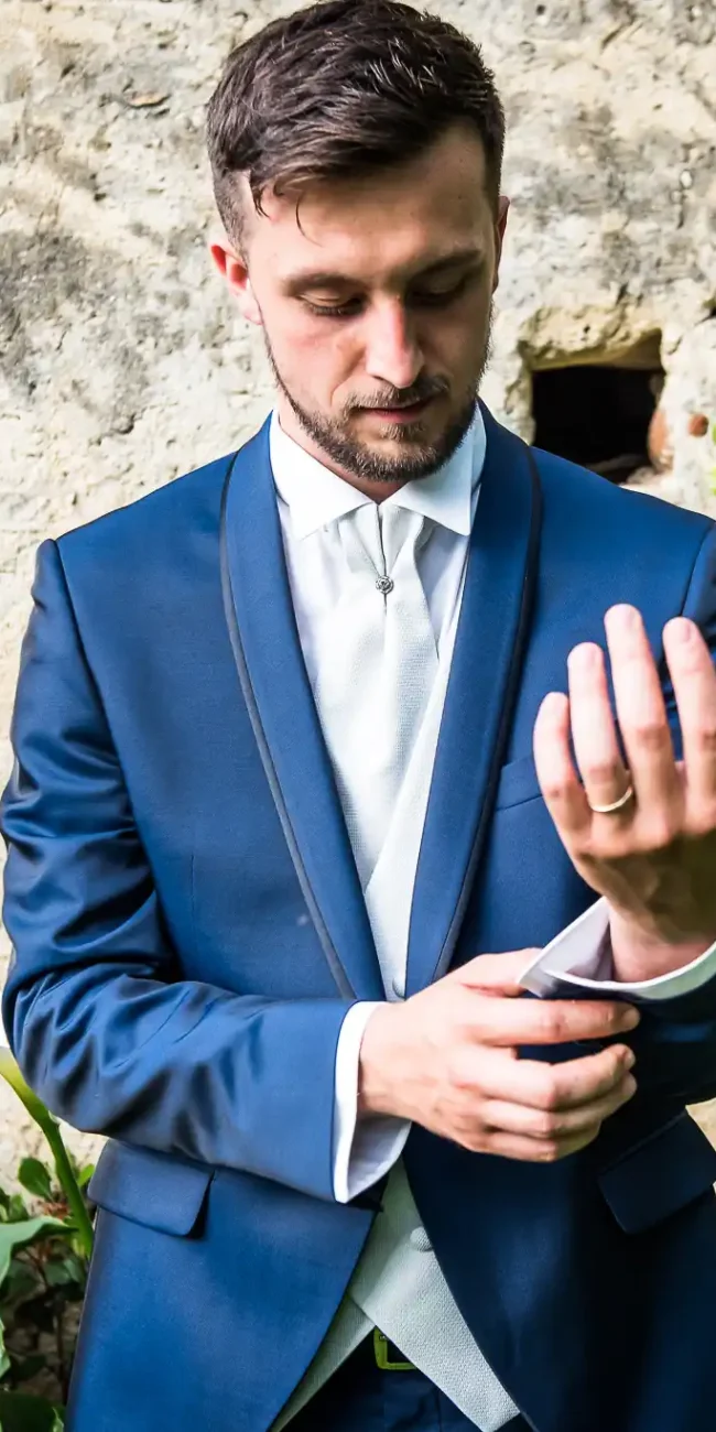 Foto matrimoniale di uno sposo in abito blu che sistema il polsino della camicia, con uno sfondo di muro di pietra e piante verdi, parte di un reportage di matrimonio.