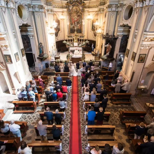 Reportage di matrimonio che cattura una vista panoramica della cerimonia in chiesa, con la coppia al centro di un tappeto rosso e gli invitati seduti ai lati.