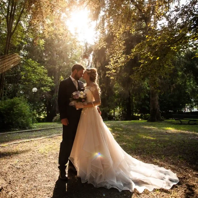 Una coppia di sposi si abbraccia teneramente sotto un albero in un parco illuminato dal sole al tramonto, con la sposa in un elegante abito bianco e lo sposo in un completo scuro, bouquet in mano, foto matrimoniali, reportage di matrimonio.