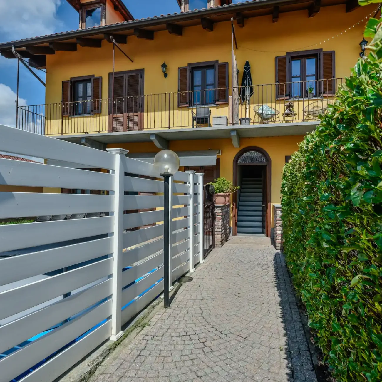 Foto immobili di un balcone esterno con ringhiera in ferro, vista su un giardino e edificio giallo, catturata da un fotografo per agenzia immobiliare.