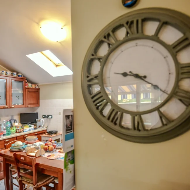 Foto immobili di un'altra vista della cucina con orologio a parete e tavolo apparecchiato, fotografata da un fotografo per agenzia immobiliare.