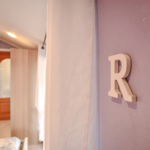 Foto immobili di una stanza con parete viola e lettera "R" decorativa, con tenda bianca e porte in legno, catturata da un fotografo per agenzia immobiliare.