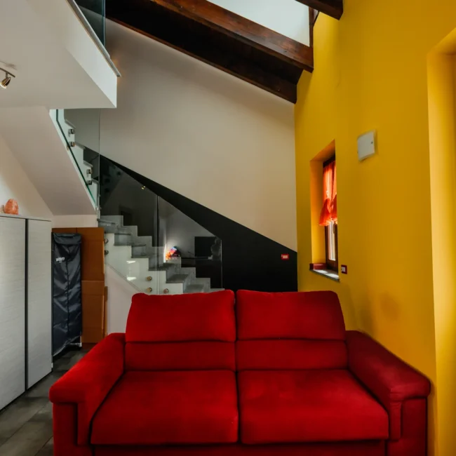 Foto immobili di un soggiorno con divano rosso e pareti gialle, con scala a vista e dettagli accoglienti, realizzata da un fotografo per agenzia immobiliare.