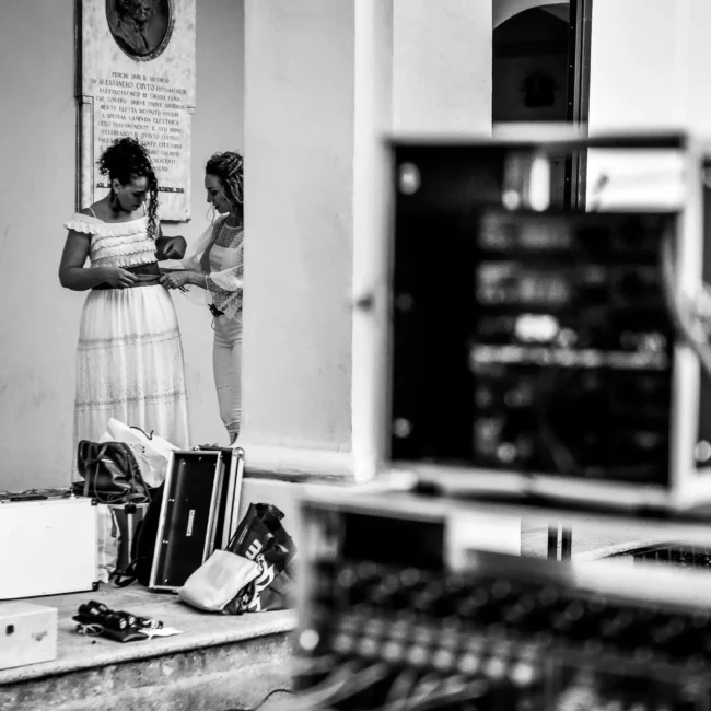 fotografo eventi Torino: Tre donne in abiti bianchi che si preparano per un evento, con attrezzature audio sullo sfondo.