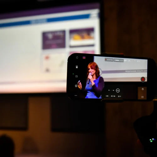 otografo eventi Torino: Una persona che registra un discorso con uno smartphone su un treppiede, con un presentatore sullo sfondo e uno schermo.