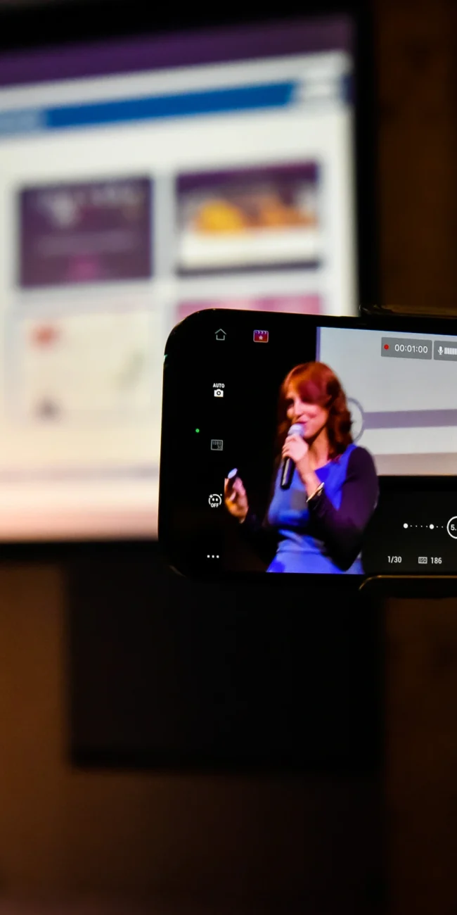 otografo eventi Torino: Una persona che registra un discorso con uno smartphone su un treppiede, con un presentatore sullo sfondo e uno schermo.