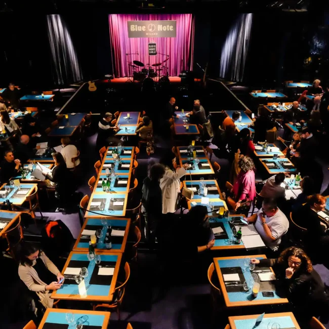 fotografo eventi Torino: Una vista dall'alto di una sala da pranzo del Blue Note Milano, con tavoli apparecchiati e un palco illuminato.