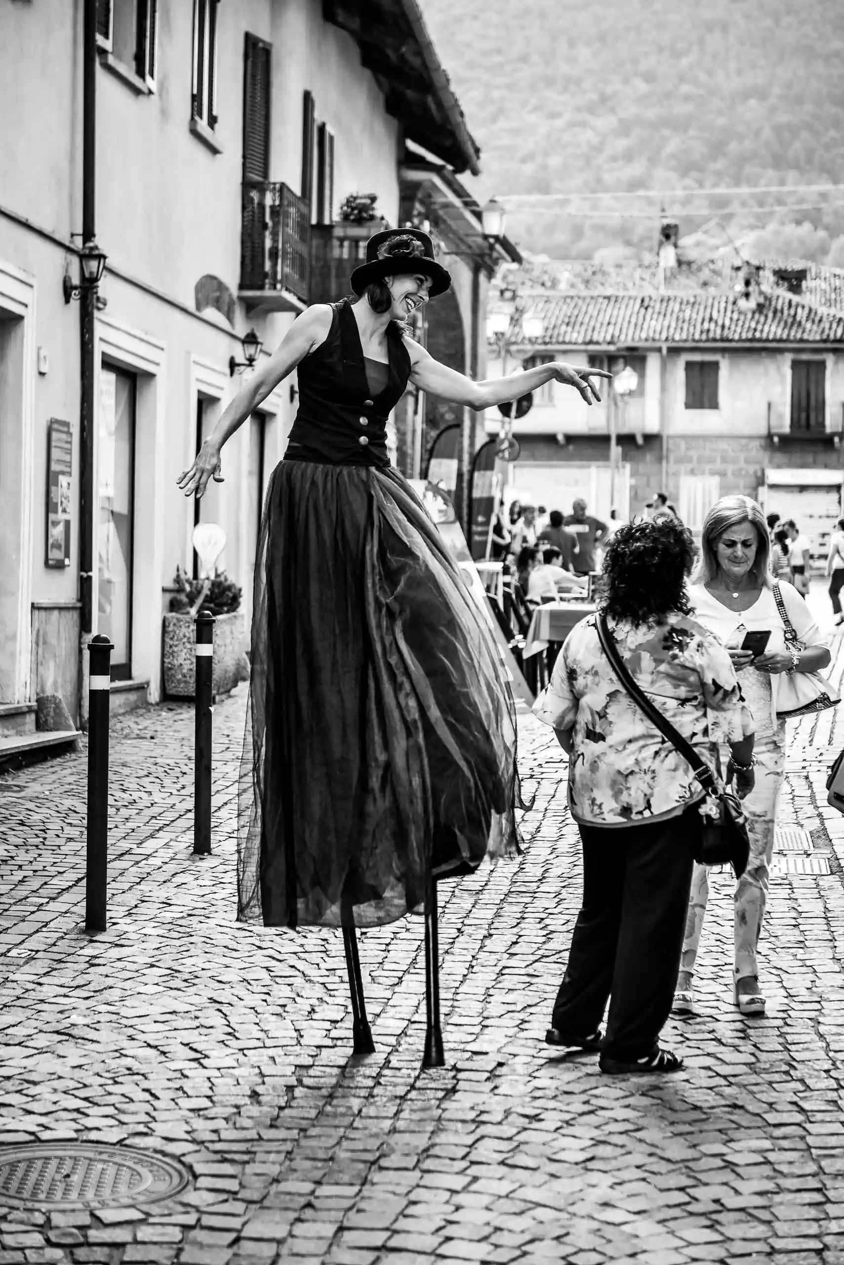 fotografo eventi Torino: Artista su trampoli in un vicolo acciottolato, in bianco e nero, interagisce con una persona che guarda il telefono.
