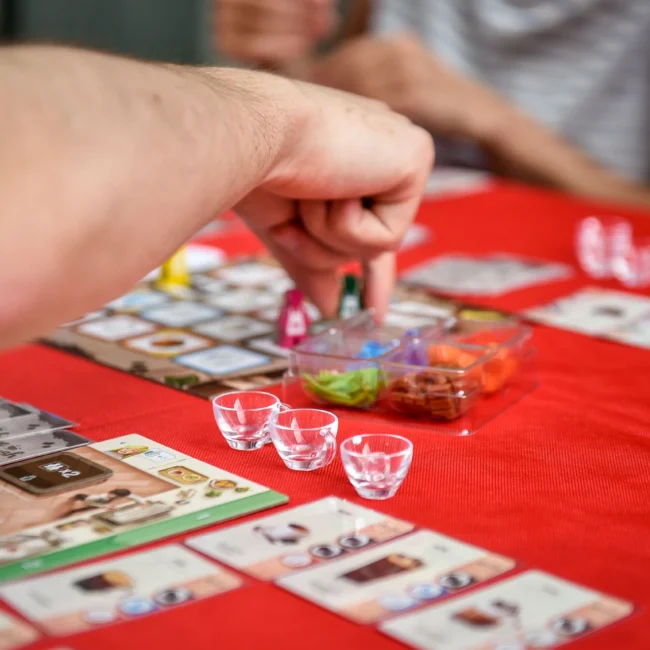 fotografo eventi Torino: Tavolo da gioco con carte e pedine, una mano che muove un pezzo, tovaglia rossa e bicchieri trasparenti.