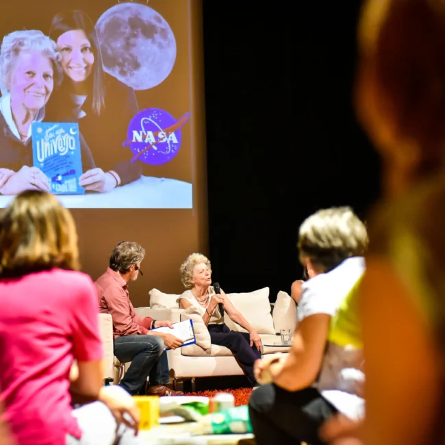 Scena di un evento con un grande schermo che mostra due donne, una che tiene un libro e una medaglia, con il logo NASA visibile.