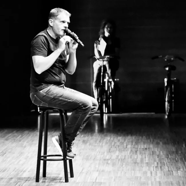 Fotografo eventi Torino riprende un Stefano massini che si esibisce su un palco con biciclette e persone sullo sfondo in bianco e nero.