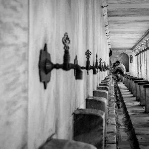 Corridoio storico con fontanelle ornamentali in pietra e un uomo seduto, catturato in bianco e nero, perfetto per viaggi per fotografi interessati ad architettura e dettagli culturali.